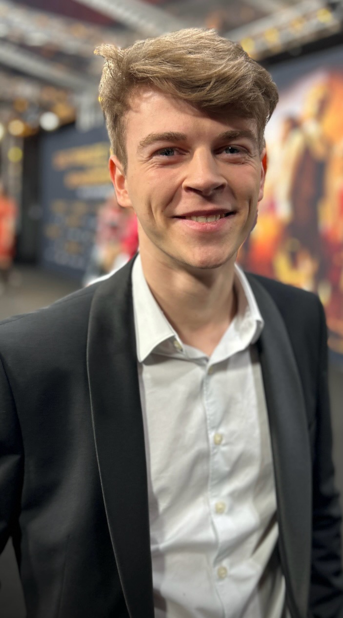Junger Mann lächelt in schwarzem Anzug und weißem Hemd auf rotem Teppich.