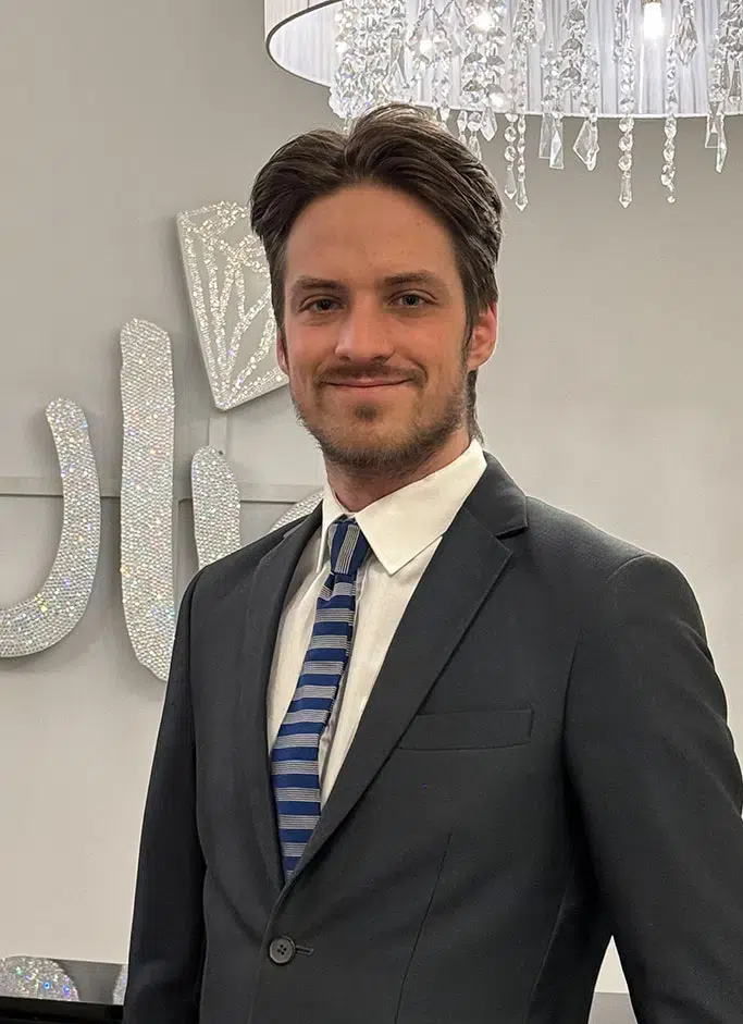 Eine Person im Anzug lächelt höflich; im Hintergrund ein Kronleuchter und glitzernde Dekorationen.