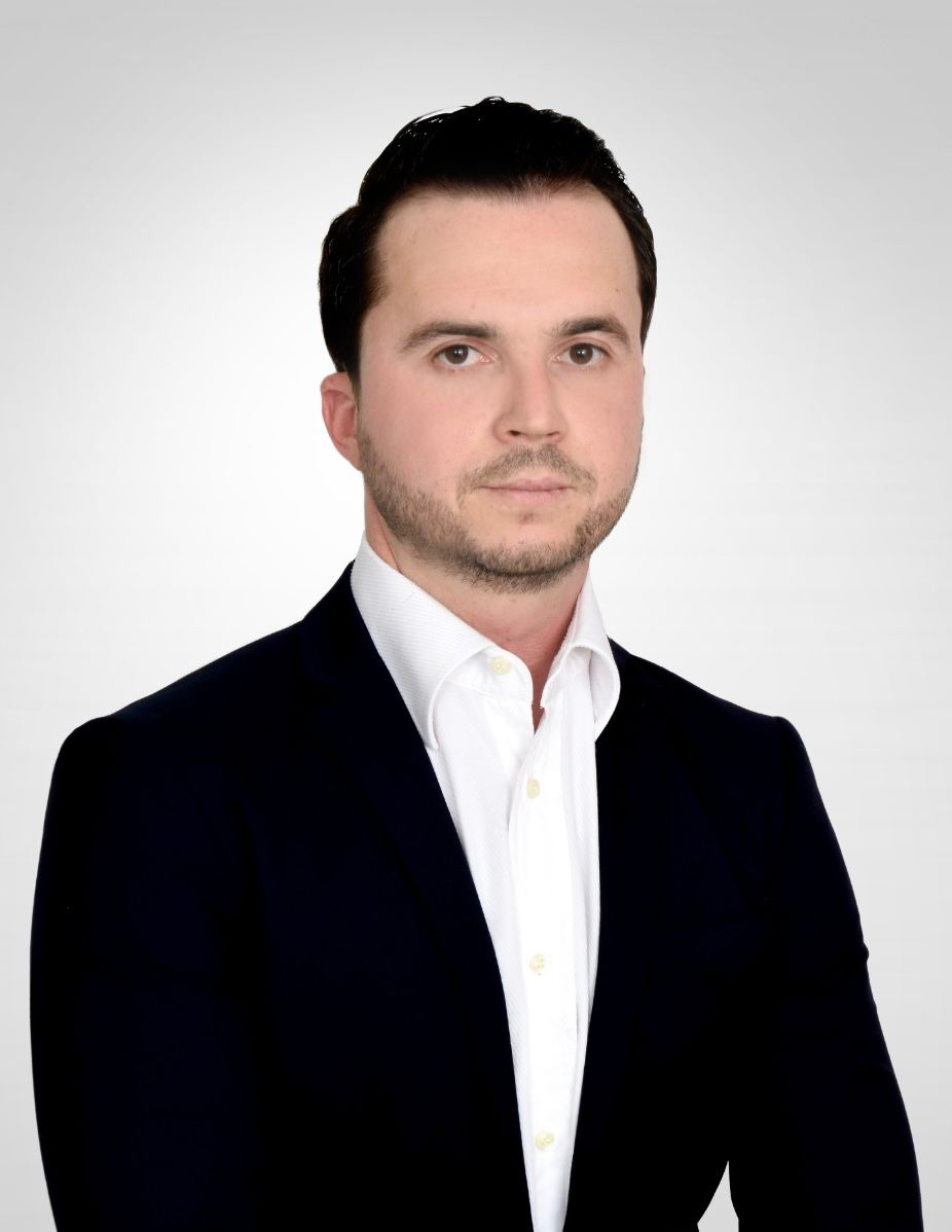 Ein Mann mit dunklem Haar und Anzug, ernster Blick, weißes Hemd, neutraler Hintergrund.
