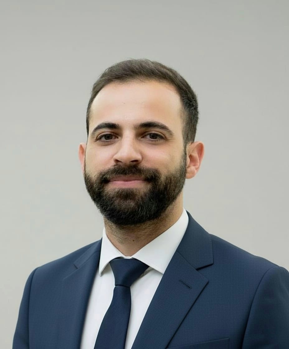 Ein Mann mit Bart und dunklem Haar lächelt dezent im blauen Anzug und weißem Hemd